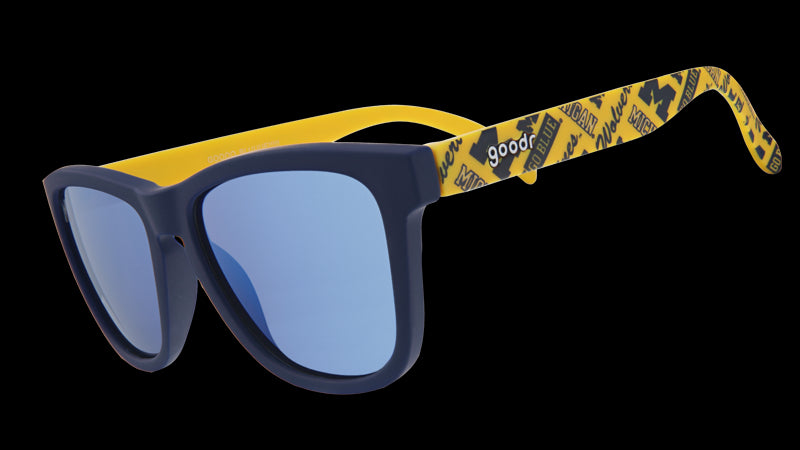A pair of "goodr" sunglasses with navy-blue frames and reflective blue lenses. The arms of the sunglasses are bright yellow and feature University of Michigan "M" logos and text that reads "Go Blue" in a repeating pattern.