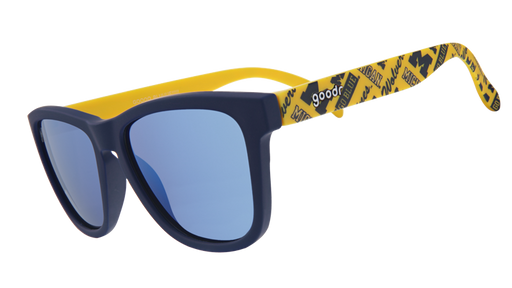 A pair of "goodr" sunglasses with navy-blue frames and reflective blue lenses. The arms of the sunglasses are bright yellow and feature University of Michigan "M" logos and text that reads "Go Blue" in a repeating pattern.