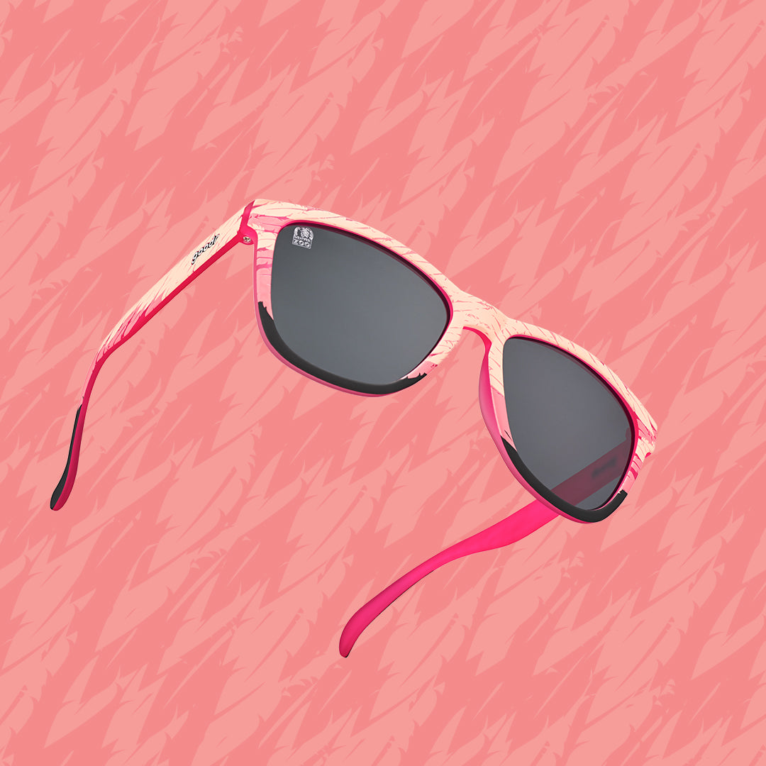 A pair of sunglasses with black lenses and a feather design in various shades of pink on the frame, on a pink background.