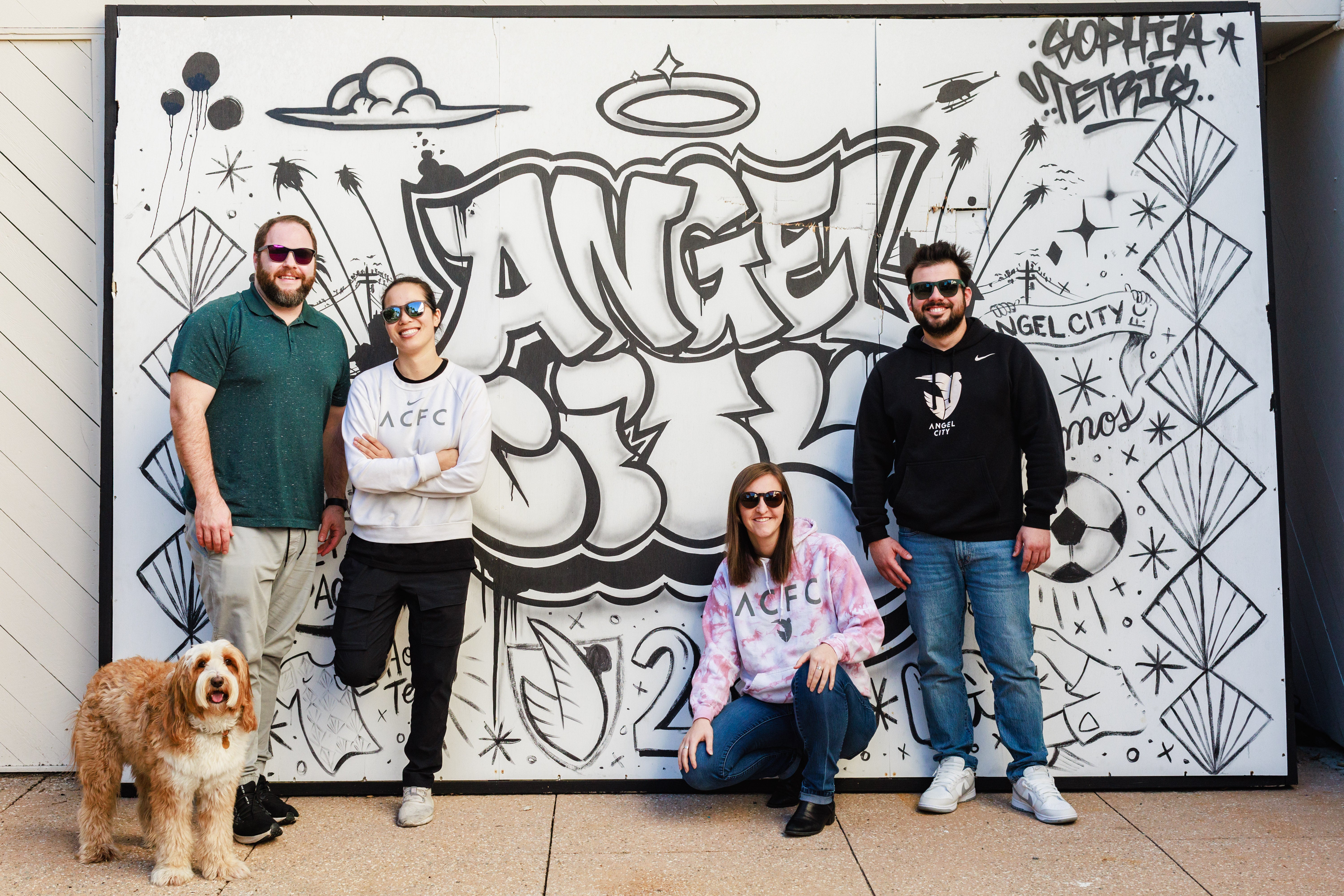 Four people wearing black sunglasses, posing in front of a graffiti wall.