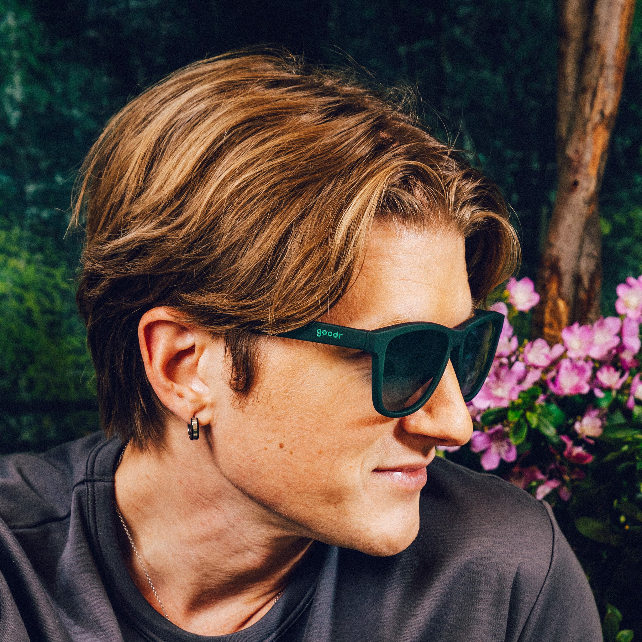 Side profile of a man wearing classic dark teal sunglasses with green gradient lenses against a nature backdrop.