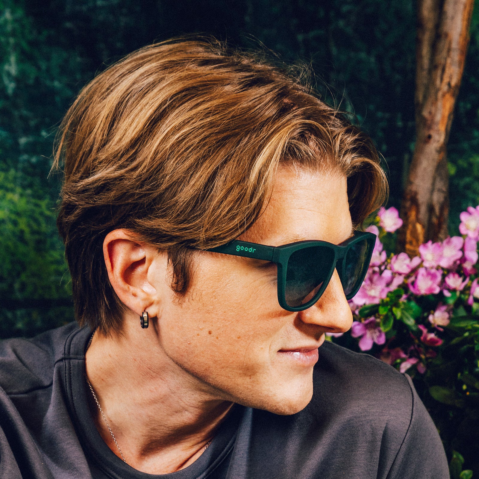 Side profile of a man wearing classic dark teal sunglasses with green gradient lenses against a nature backdrop.