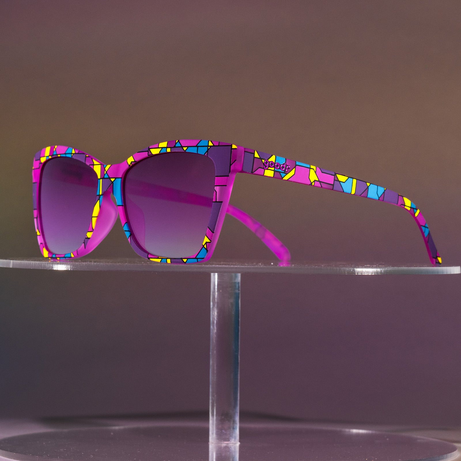 A pair of purple Pop G sunnies with a stained glass-inspired design and purple gradient lenses sits atop a clear pedestal.