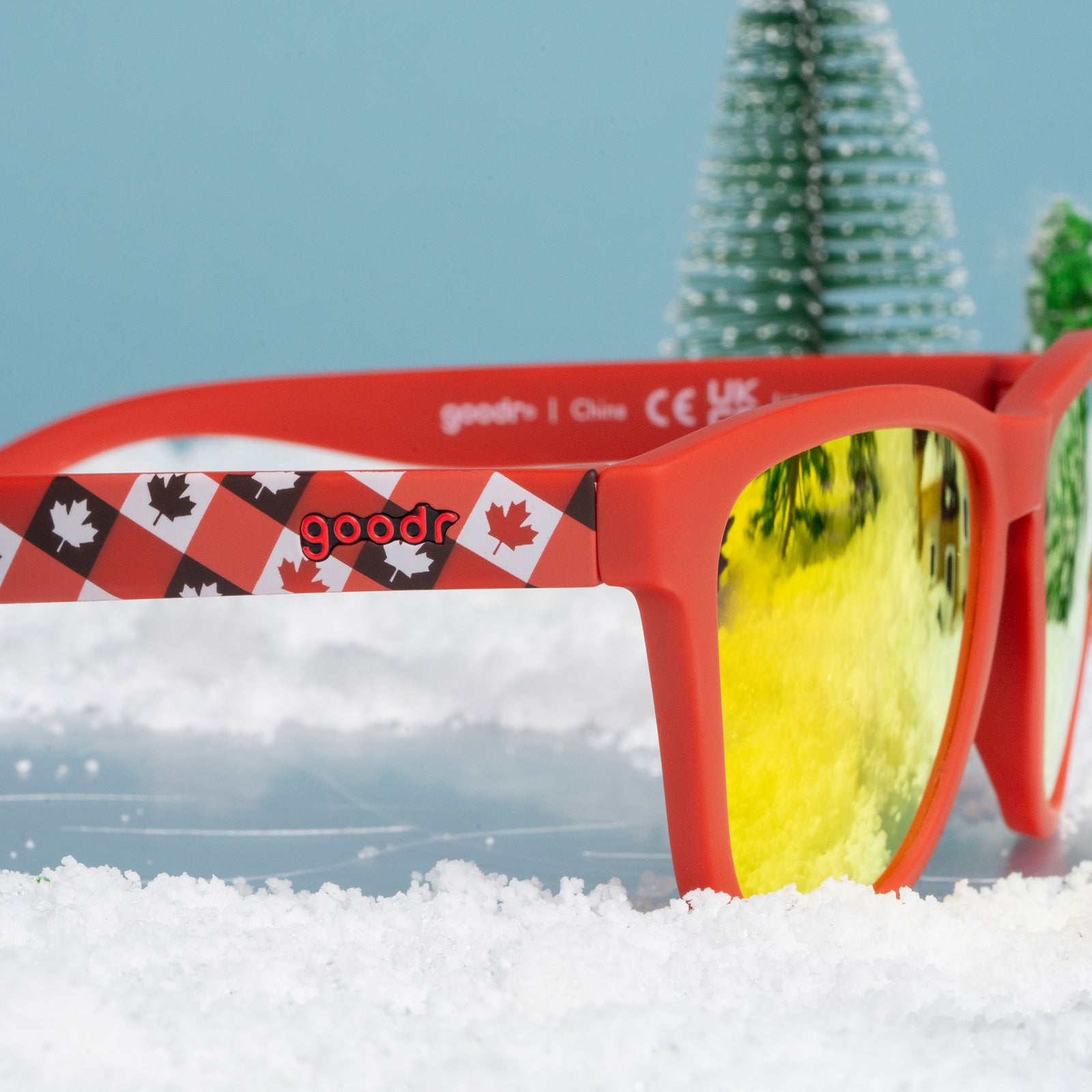Close-up side view of red maple-leaf patterned Canada sunglasses resting on snow with a wintry pine tree background.