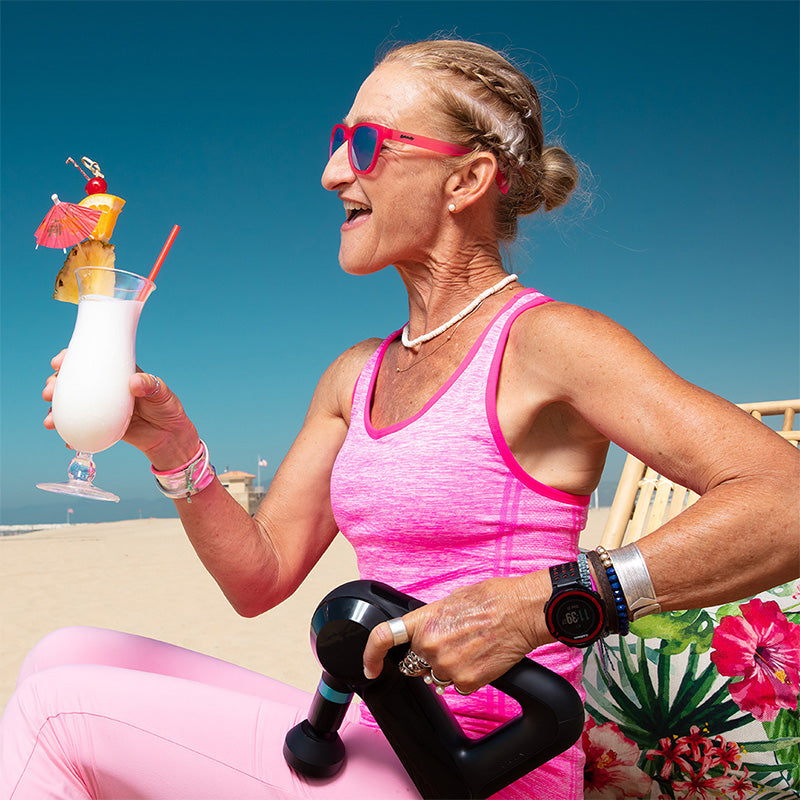 A female runner in hot pink sunglasses with teal lenses lounges on a beach with a piña colada and massage gun for her legs.