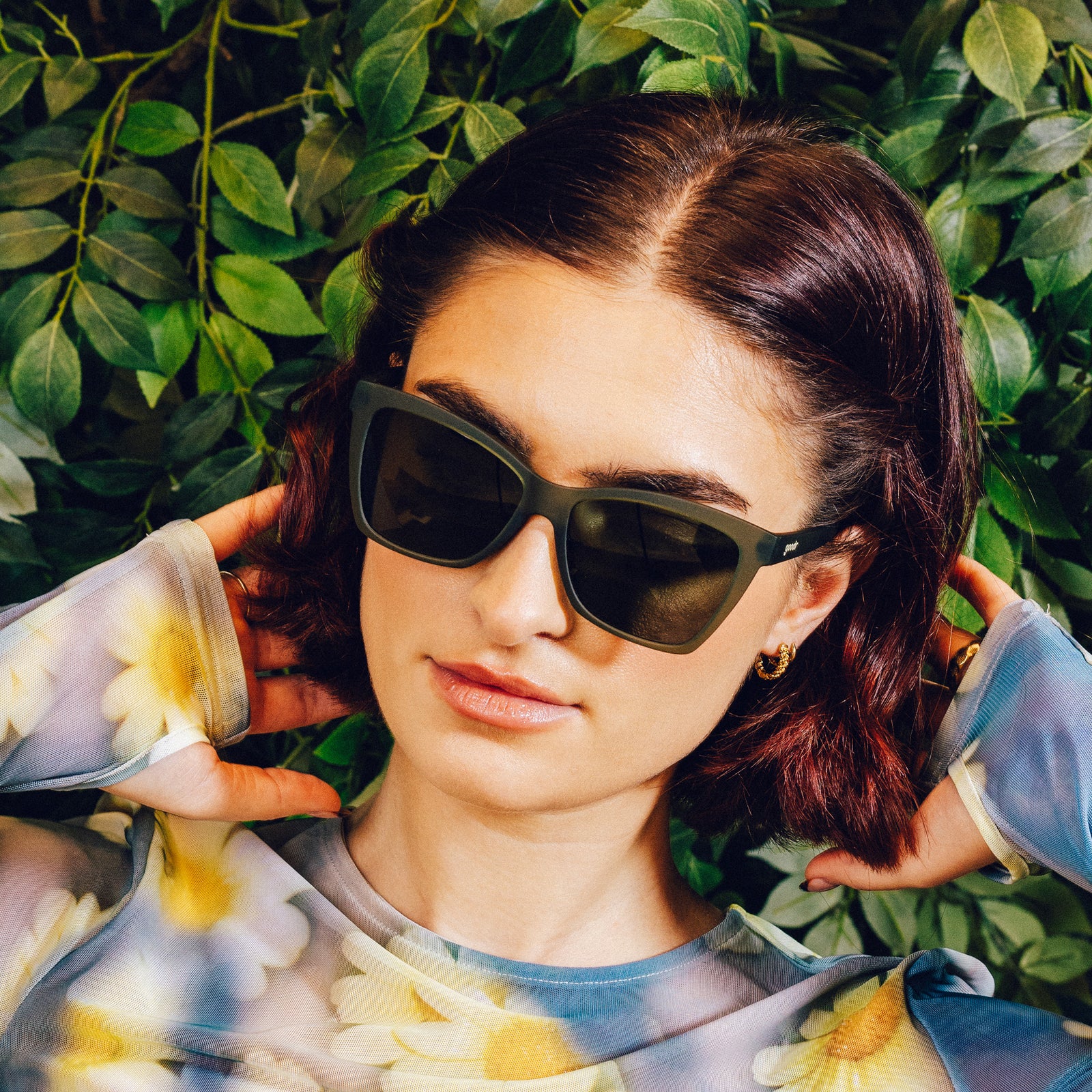 Woman poses in front of a vine backdrop while wearing translucent dark green angular sunglasses with green non-reflective lenses.
