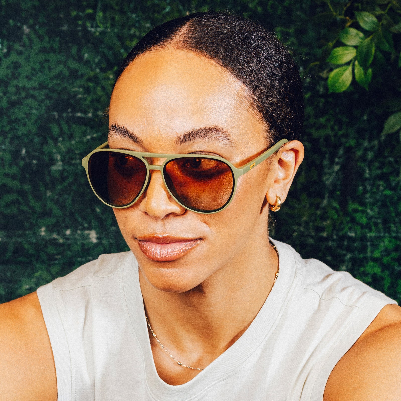 Woman sits in front of a nature backdrop while wearing green aviator sunglasses with amber gradient lenses.