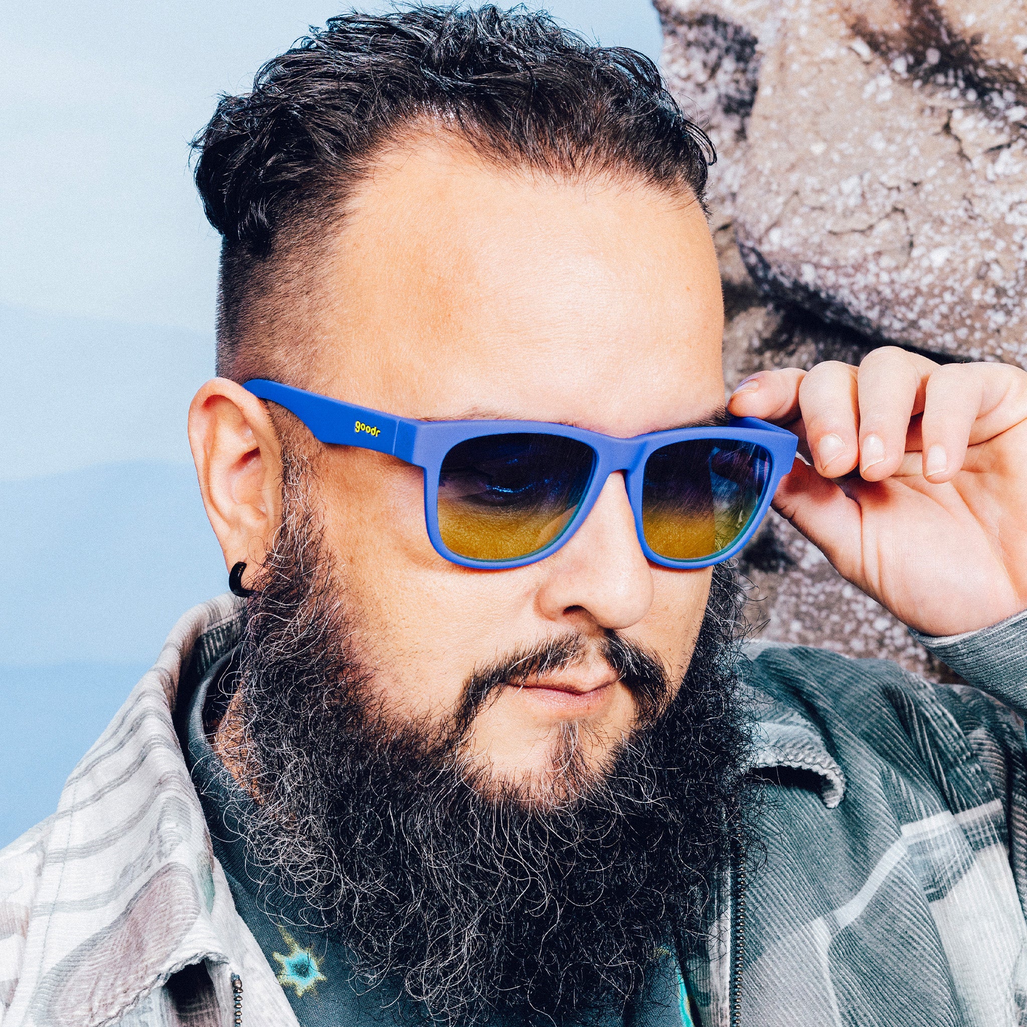 Side profile of a man posing and wearing large blue glasses with blue-yellow ocean gradient lenses in front of a mountain backdrop.