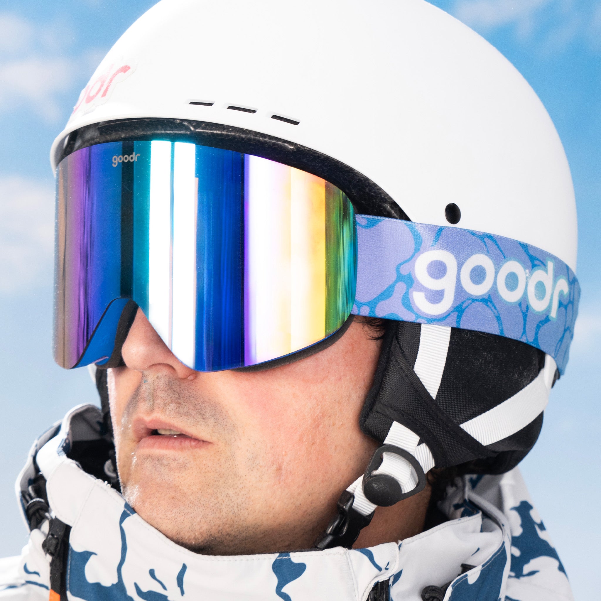 A man in a white helmet models cobalt-blue snow goggles with green reflective lenses and a blue and purple gradient strap.