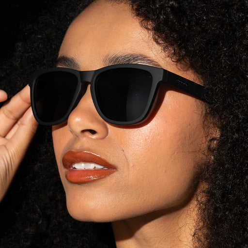 A closeup of a woman's face, wearing black square-shaped sunglasses.