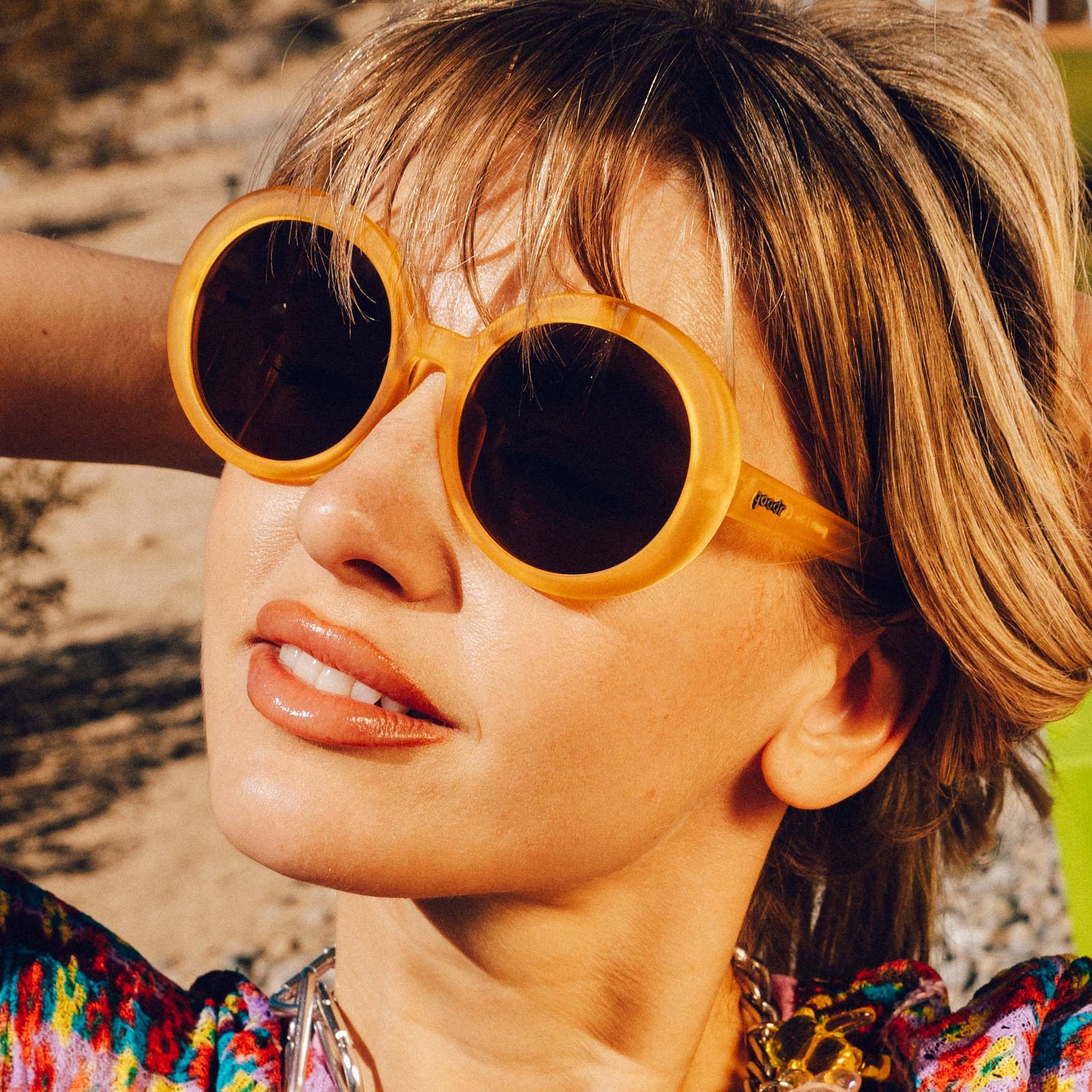 Three-quarter profile shot of a woman with medium blonde hair and glossy lips wearing a pair of oversized round orange sunglasses.