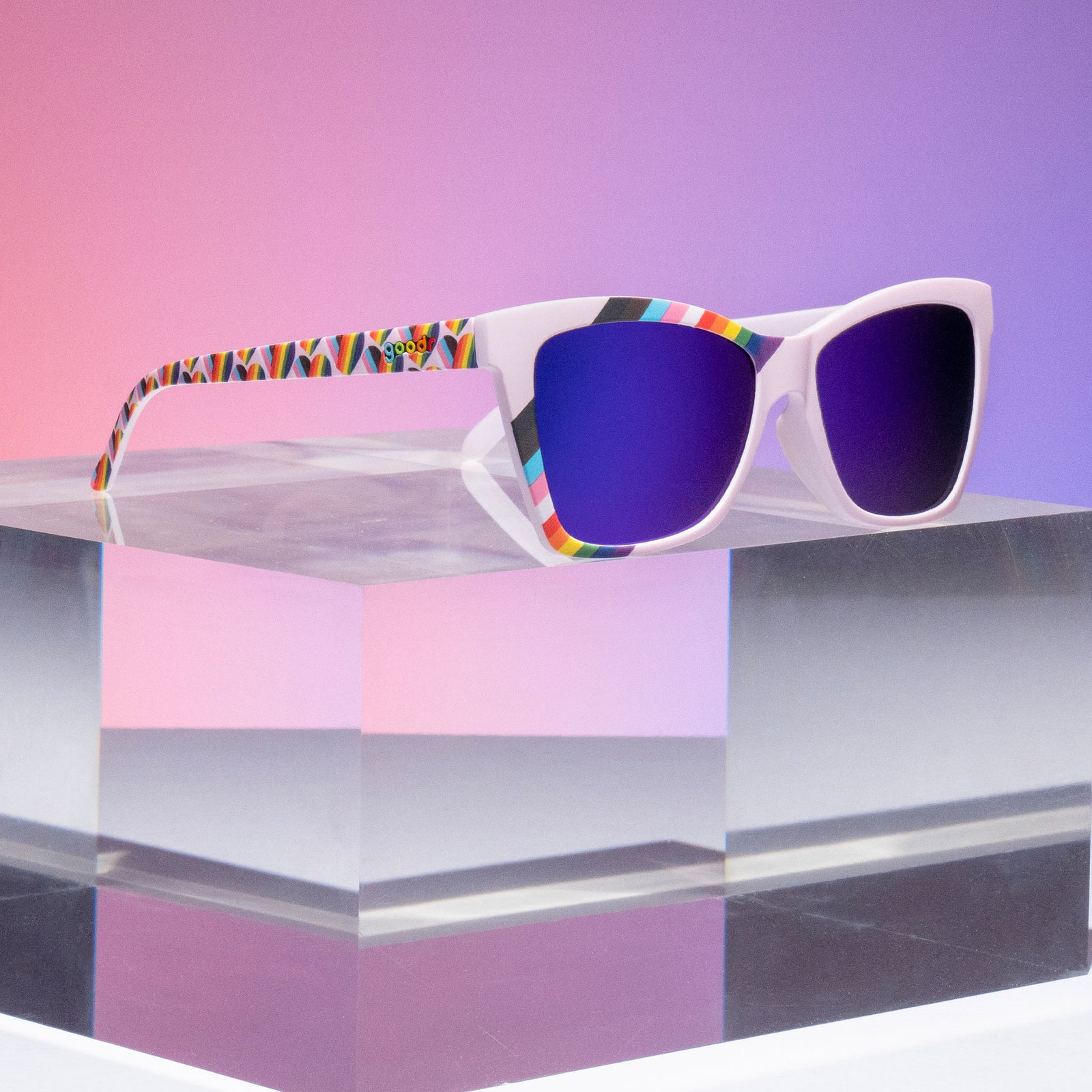 Three-quarter angle view of angled cat-eye LGBTQ Pride sunglasses with lilac frames and purple lenses on a clear podium.