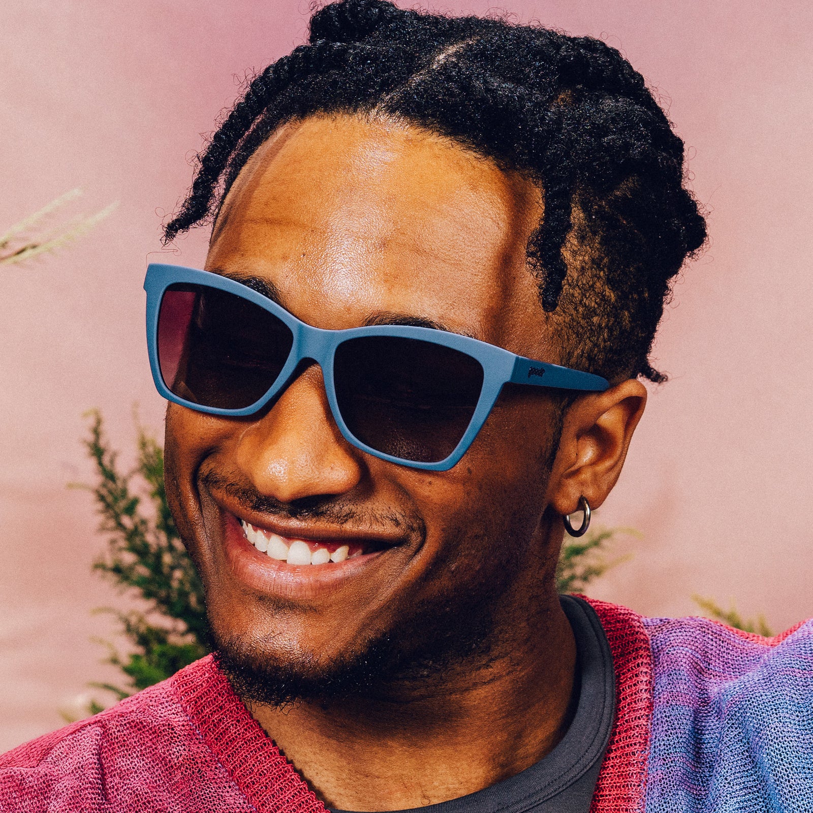 A man wearing dark purple angular sunglasses with purple gradient lenses smiles in front of a meadow backdrop.