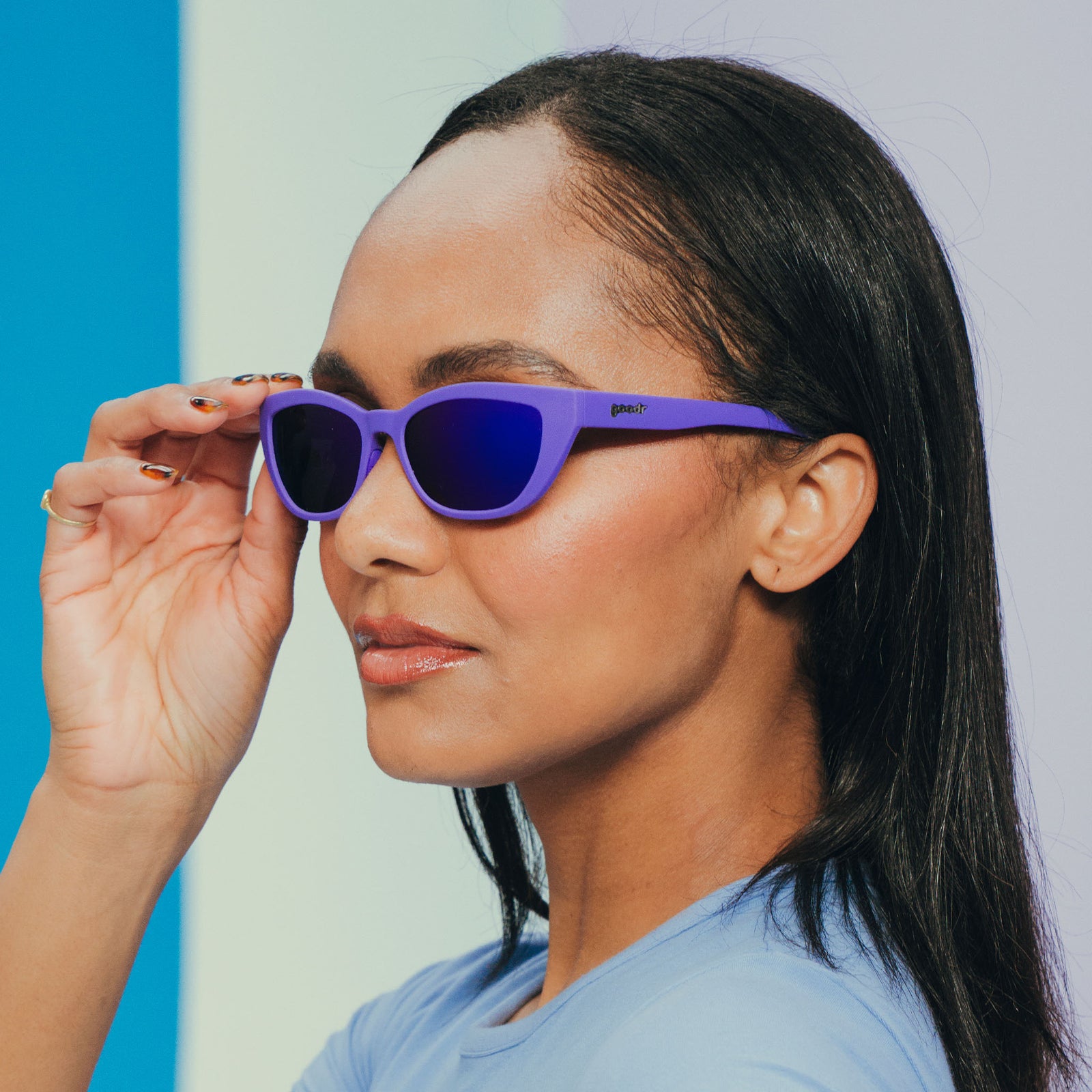 A dark-haired woman in light blue is modeling a pair of deep purple sunglasses with her hand placed on the right temple.