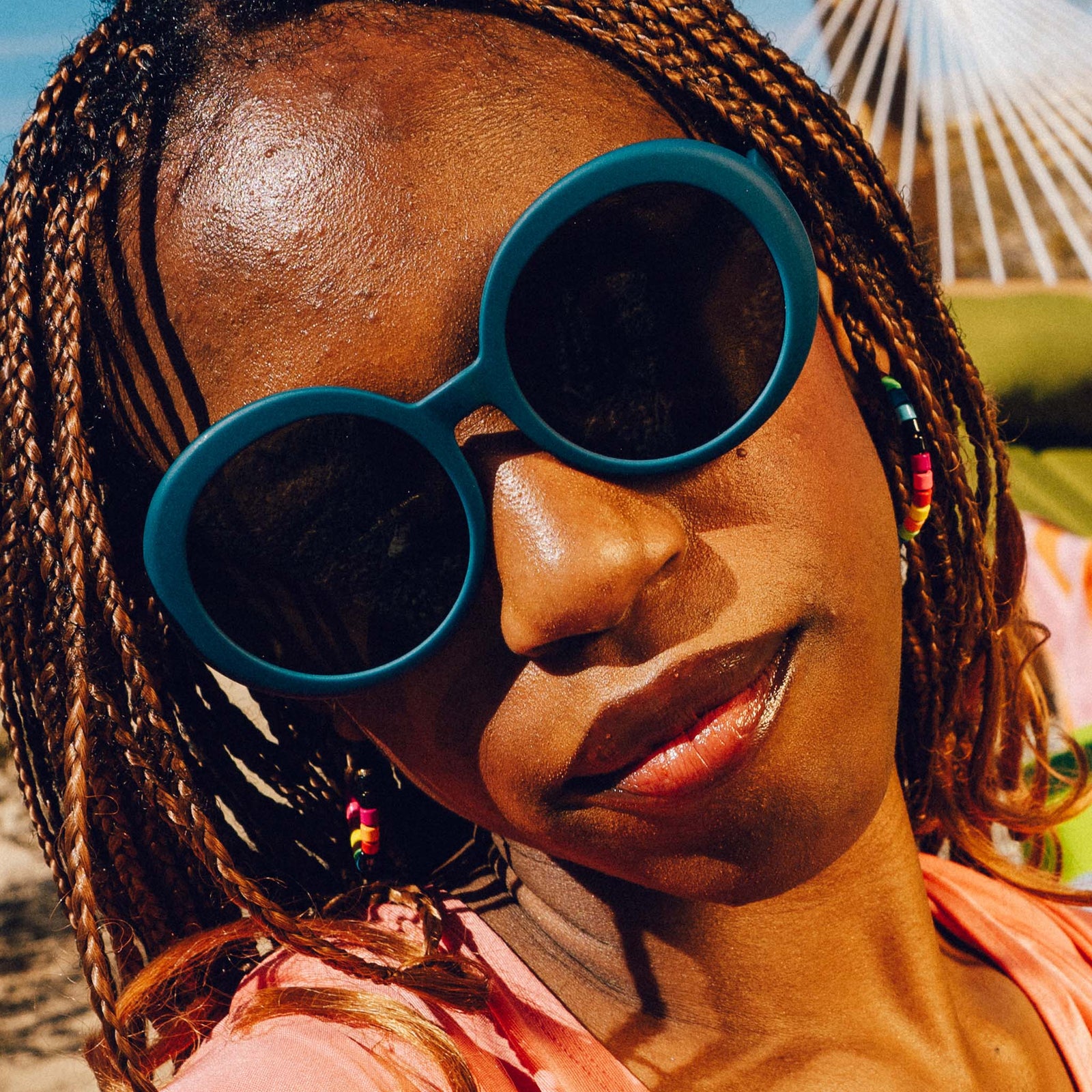 Close-up, front view of a woman's face. She has copper micro braids, colorful beaded hoop earrings, and oversized round sunglasses in deep green.