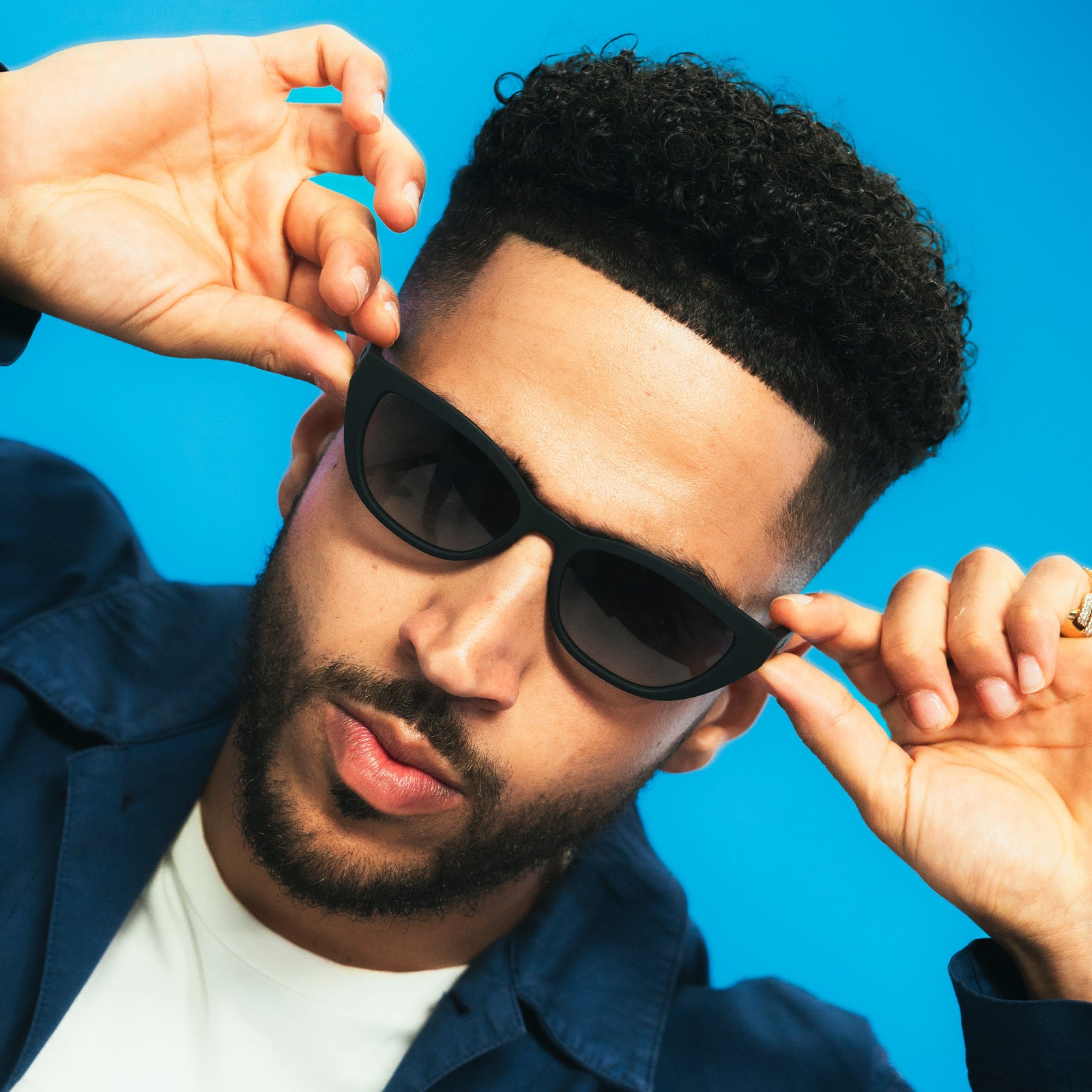 A cool dude with a fade and dark beard rocks a pair of black cat-eye sunnies, posing with his hands on the glasses' temples.