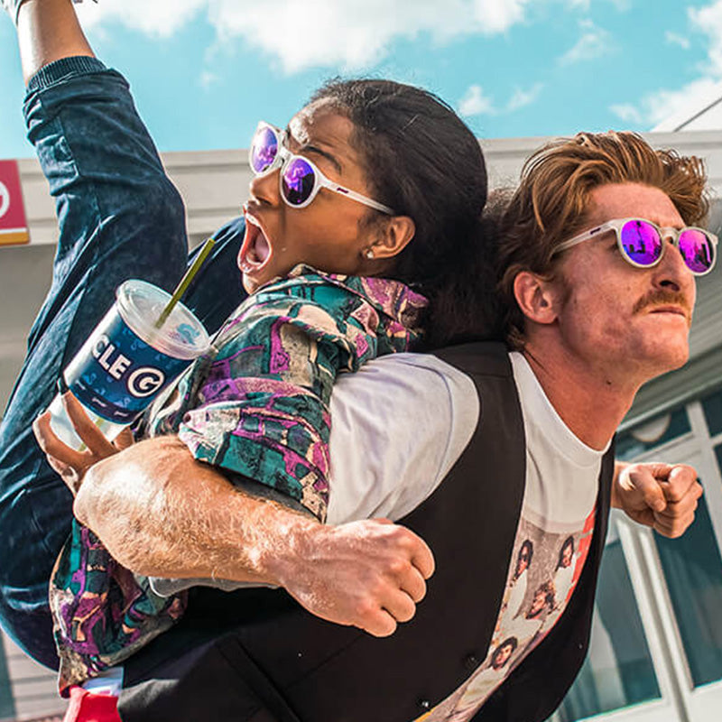 A man carries a woman sipping a gas station drink on his back, they both wear round clear sunglasses with purple lenses.