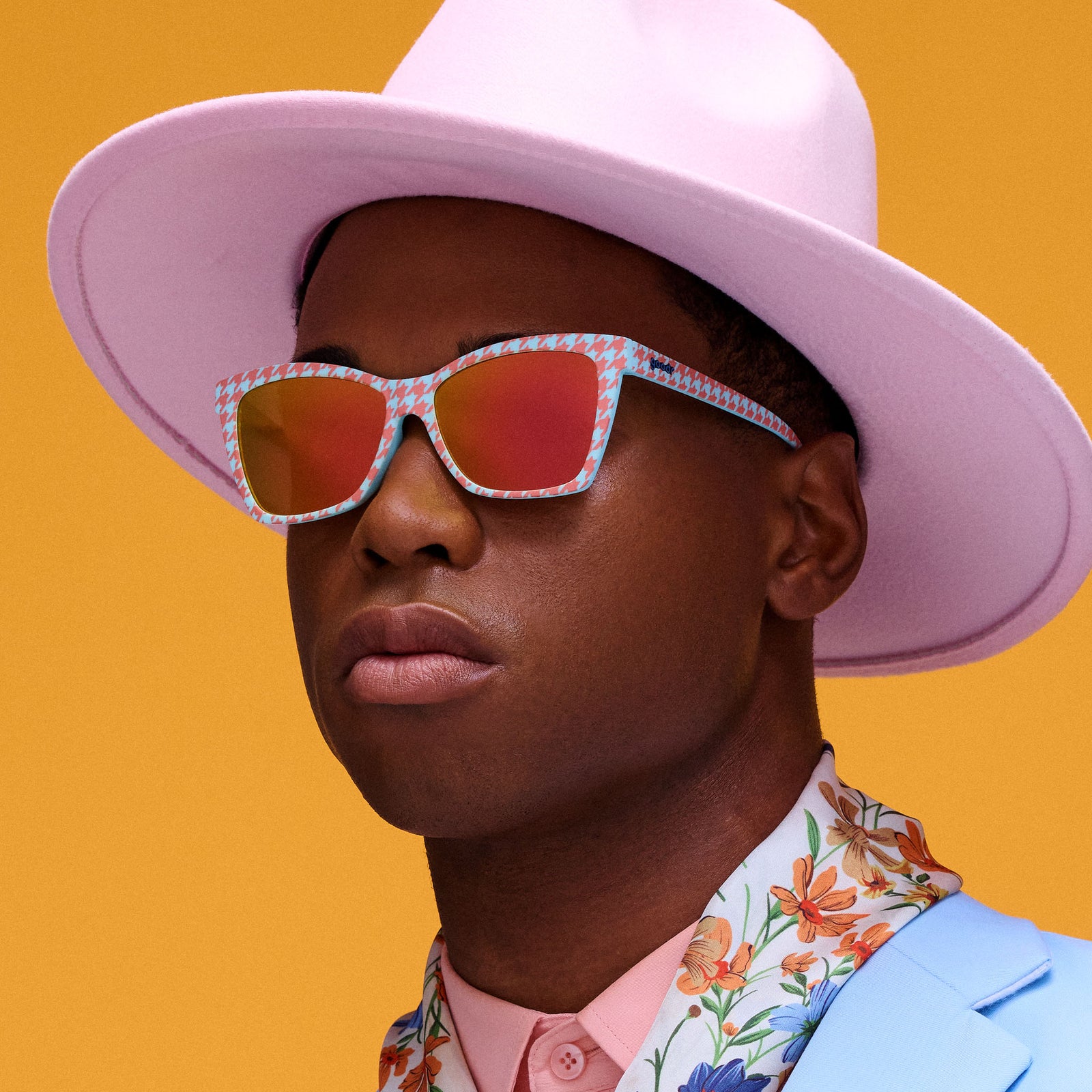 A dapper gentleman in a floral shirt, blazer, and pastel pink fedora wears blue and orange houndstooth print Pop G shades.