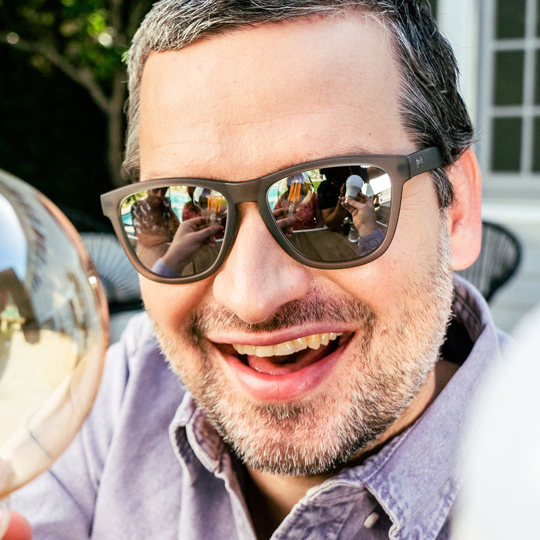 A smiling man wearing reflective sunglasses, capturing the reflections of people and outdoor surroundings, enjoying a relaxed moment in a sunny setting.
