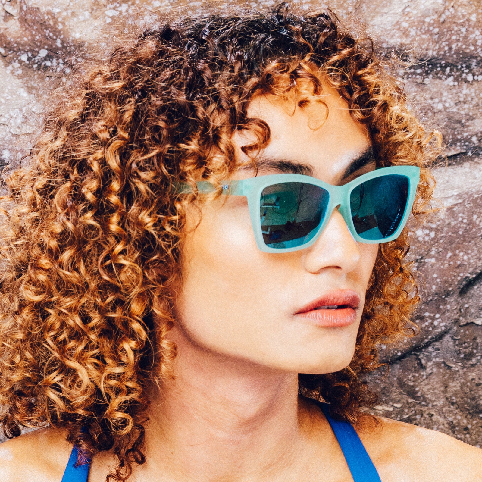 Side profile of a woman wearing translucent teal angular sunglasses with reflective teal lenses.