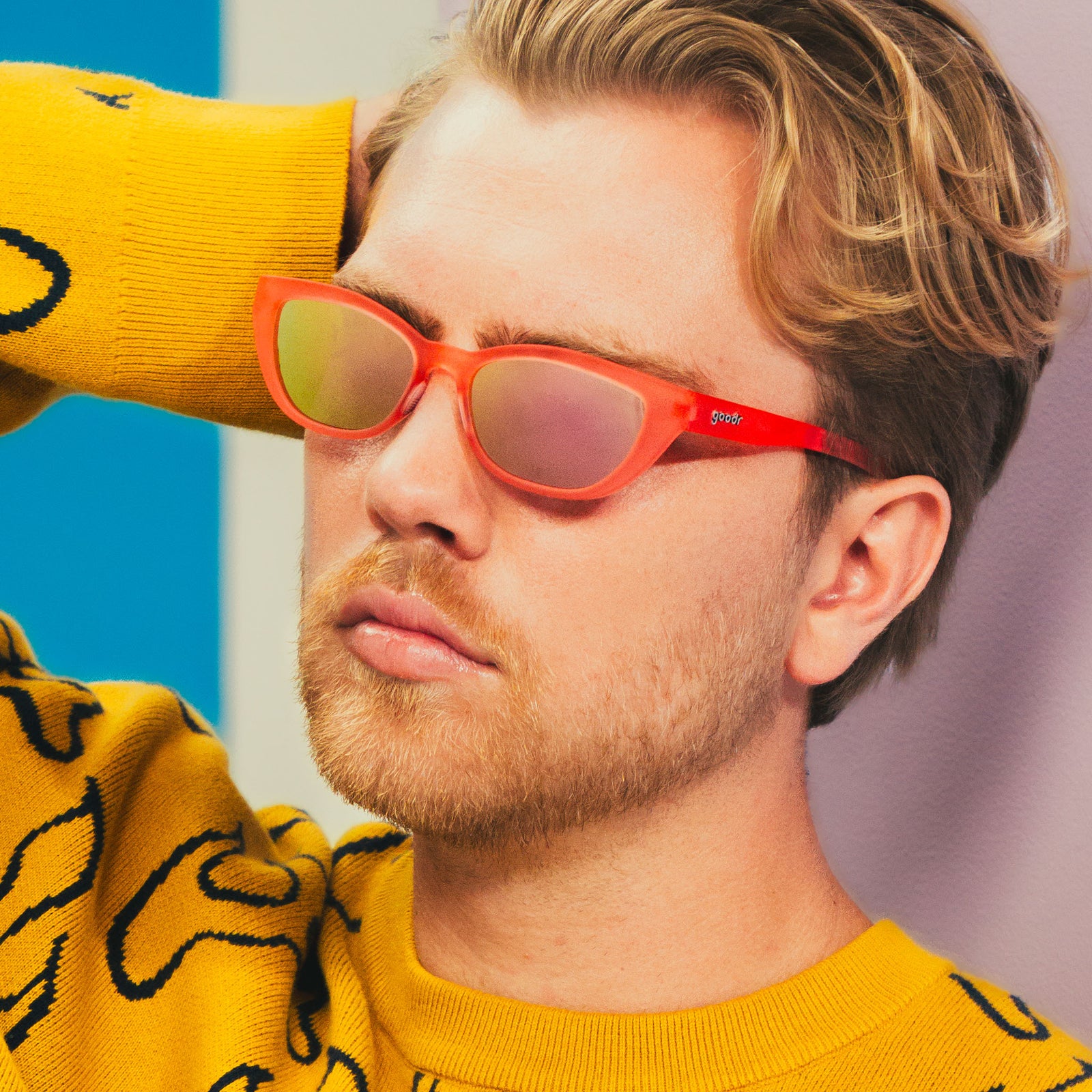 A blonde man wearing an abstract yellow sweater and translucent pink cat-eye Cheetah G frames runs his hand through his hair.