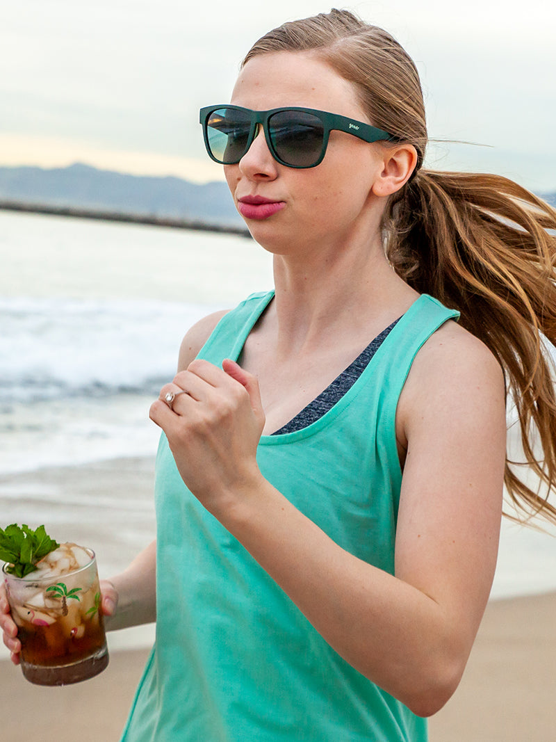 A woman running by the beach, wearing a turquoise tank top and teal sunglasses.
