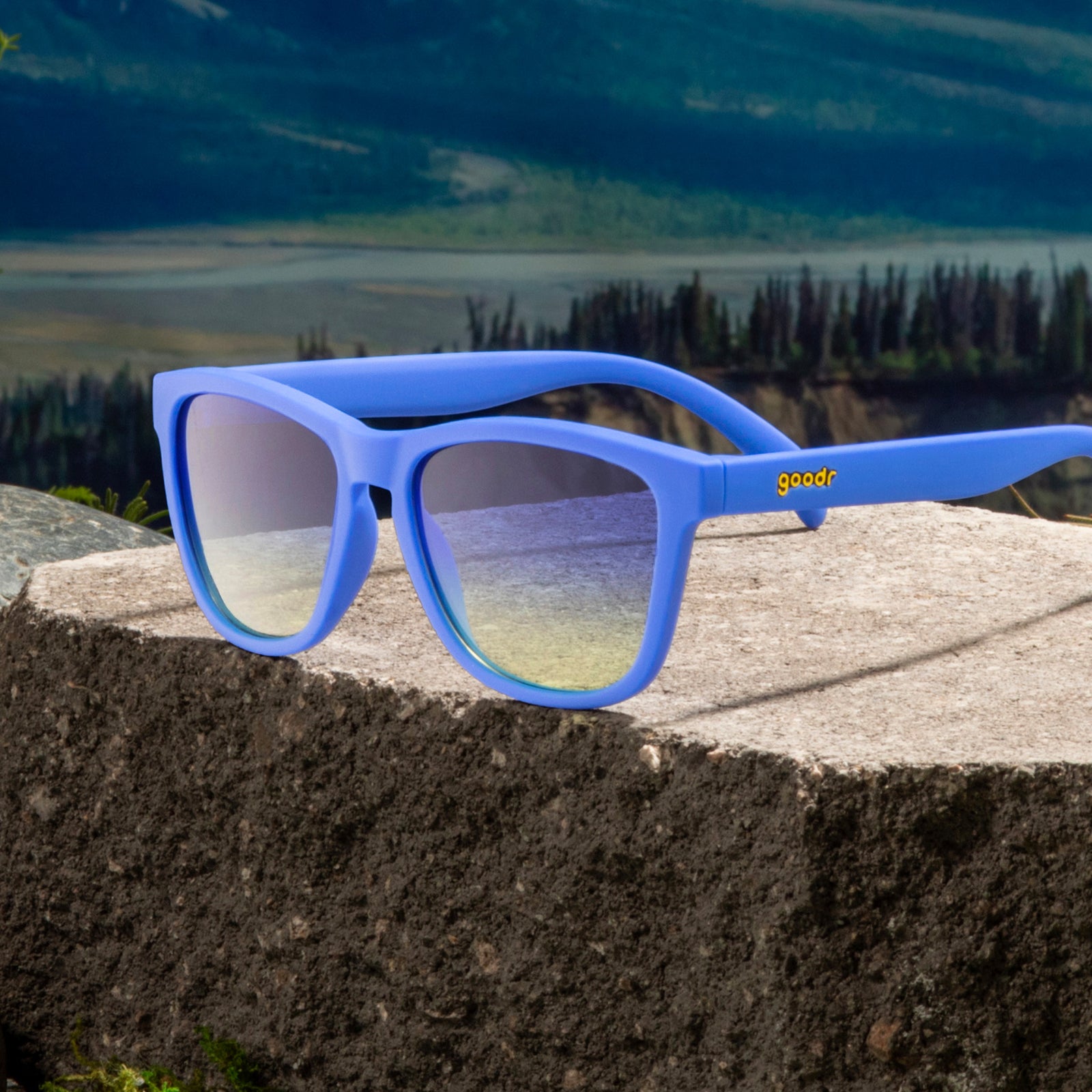 Three-quarter angle view of classic blue sunglasses with blue-yellow ocean lenses on a boulder against a mountain backdrop.