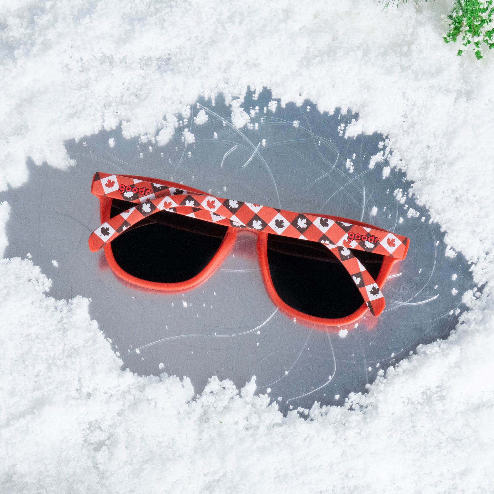 Top-down view of red maple leaf-patterned Canada sunglasses resting face down on a patch of ice surrounded by snow.