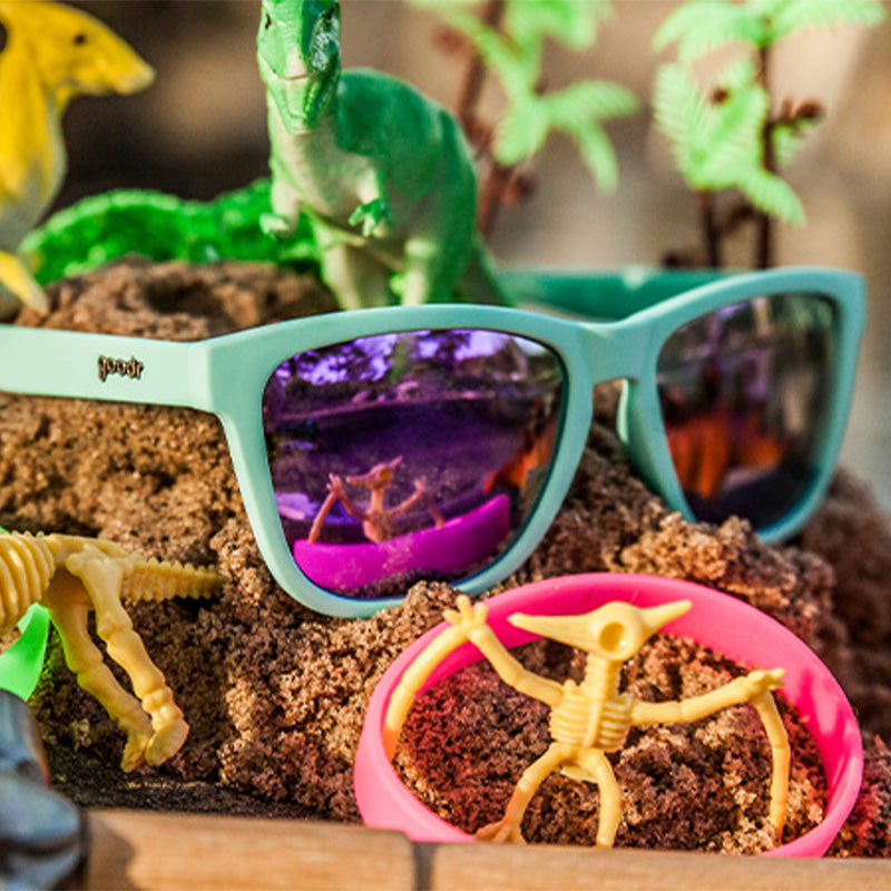 Three-quarter angle view of baby blue sunglasses with purple reflective lenses sitting on a dirt mound beside dinosaur toys.