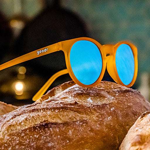 A pair of orange-framed sunglasses with blue reflective lenses is placed on top of freshly baked loaves of bread. The "goodr" logo is visible on the side of the sunglasses, and the scene has a warm, cozy feel.