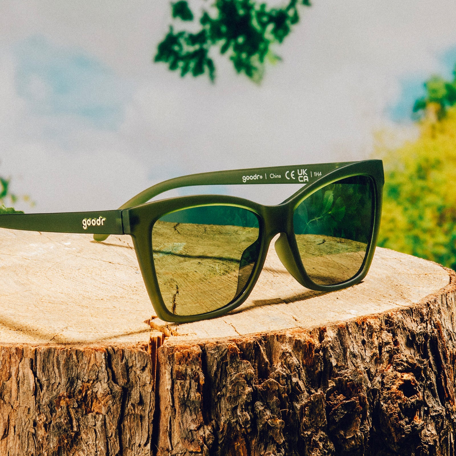 Three-quarter angle view of translucent dark green angular sunglasses with green non-reflective lenses in front of a nature backdrop.