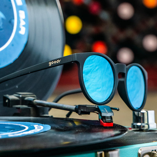 A pair of sunglasses with black, round frames and blue lenses, sitting on top of a black record player.