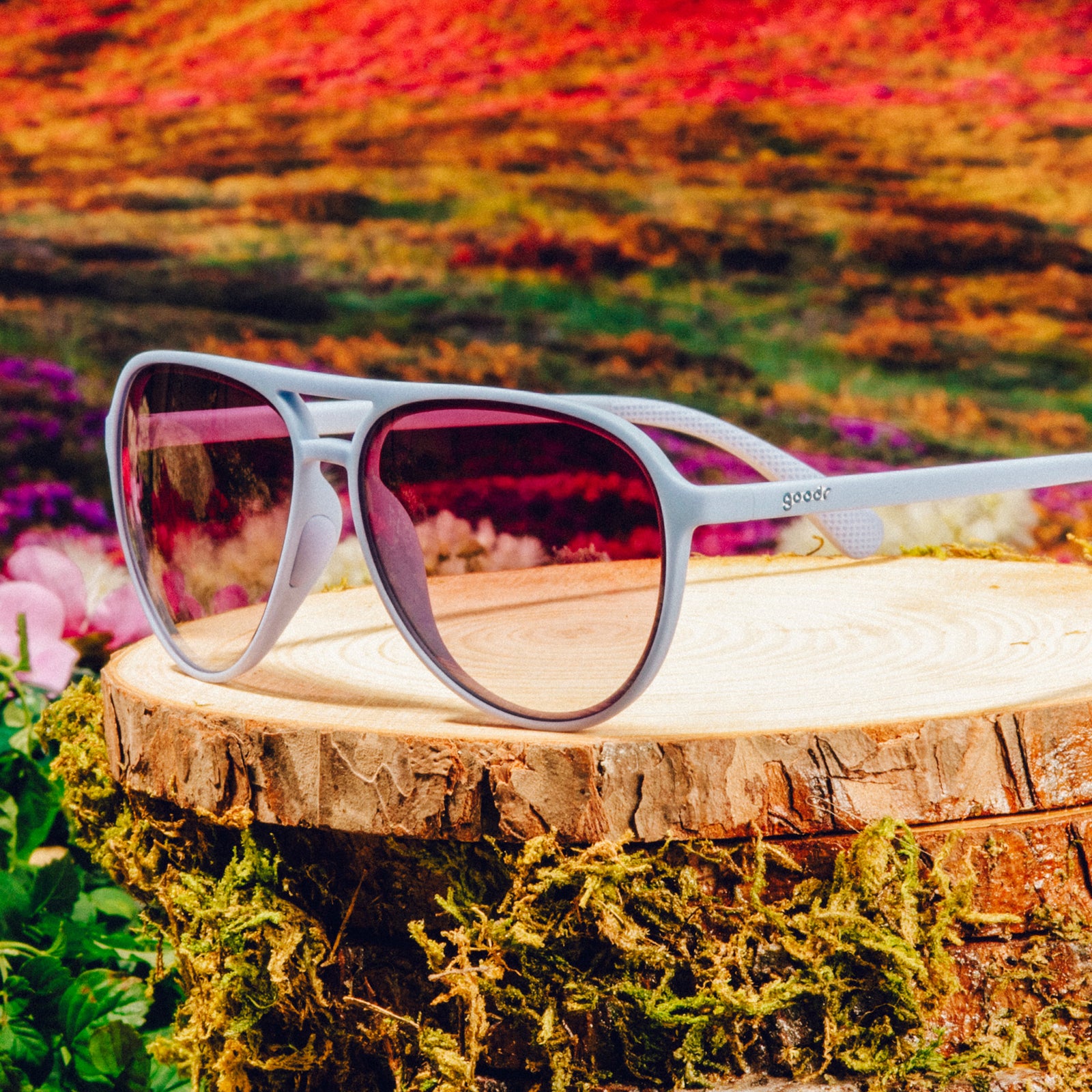 Three-quarter angle view of light blue aviator sunglasses with purple gradient lenses on a tree trunk against a meadow backdrop.