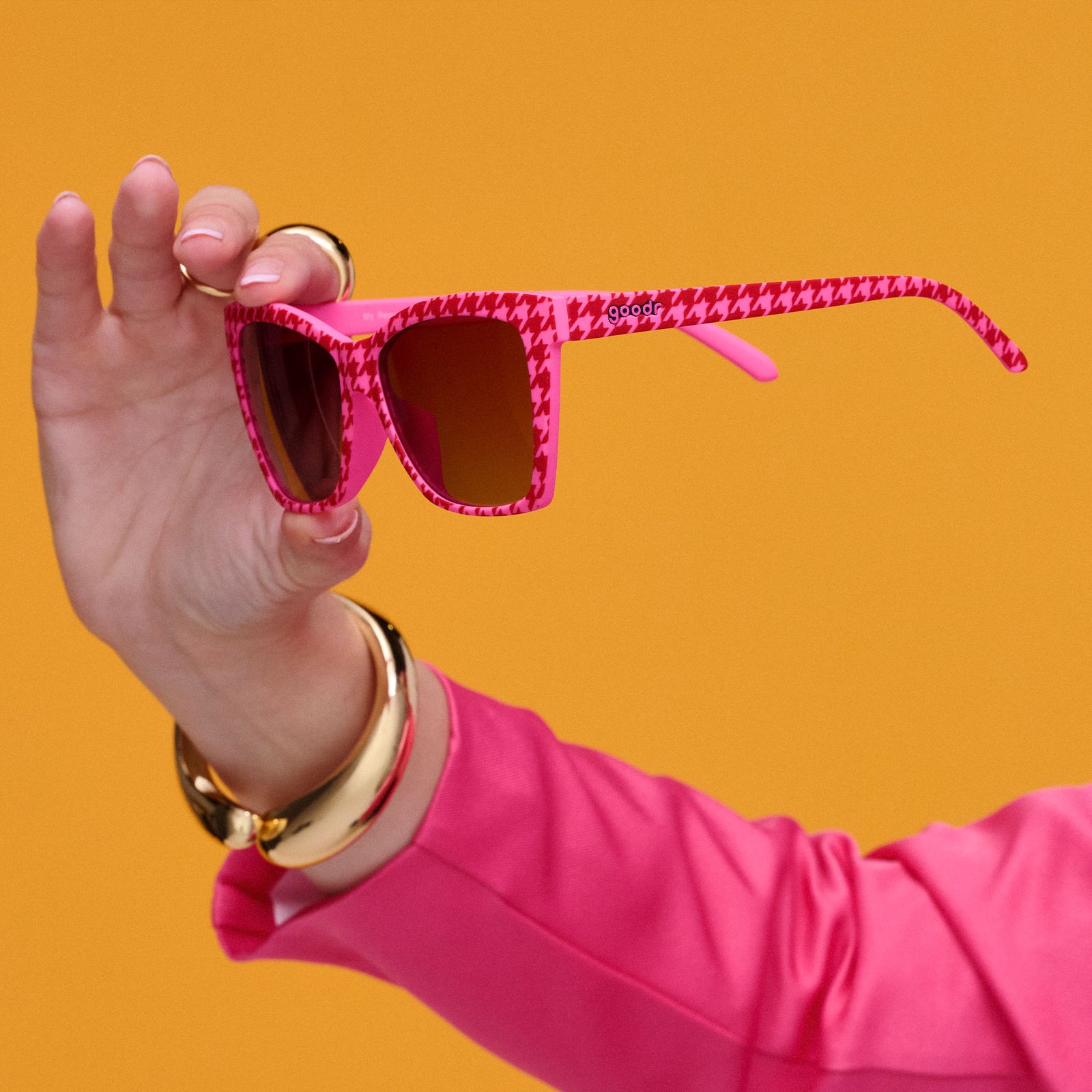 A woman’s arm in a hot pink jacket and gold jewelry holds pink and red houndstooth Pop Gs in her outstretched hand.