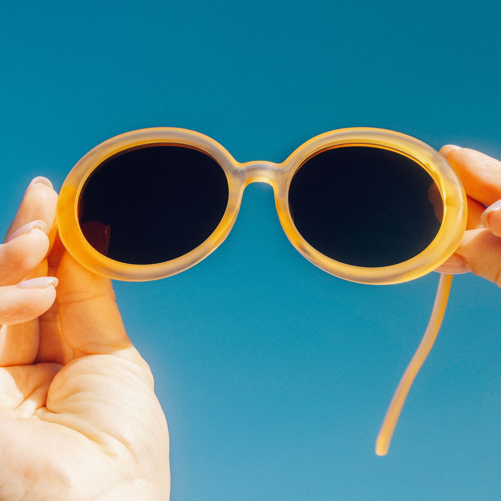 A pair of feminine, fair-skinned hands hold a pair of oversized, pale orange, round sunglasses with non-reflective lenses up to the blue sky.
