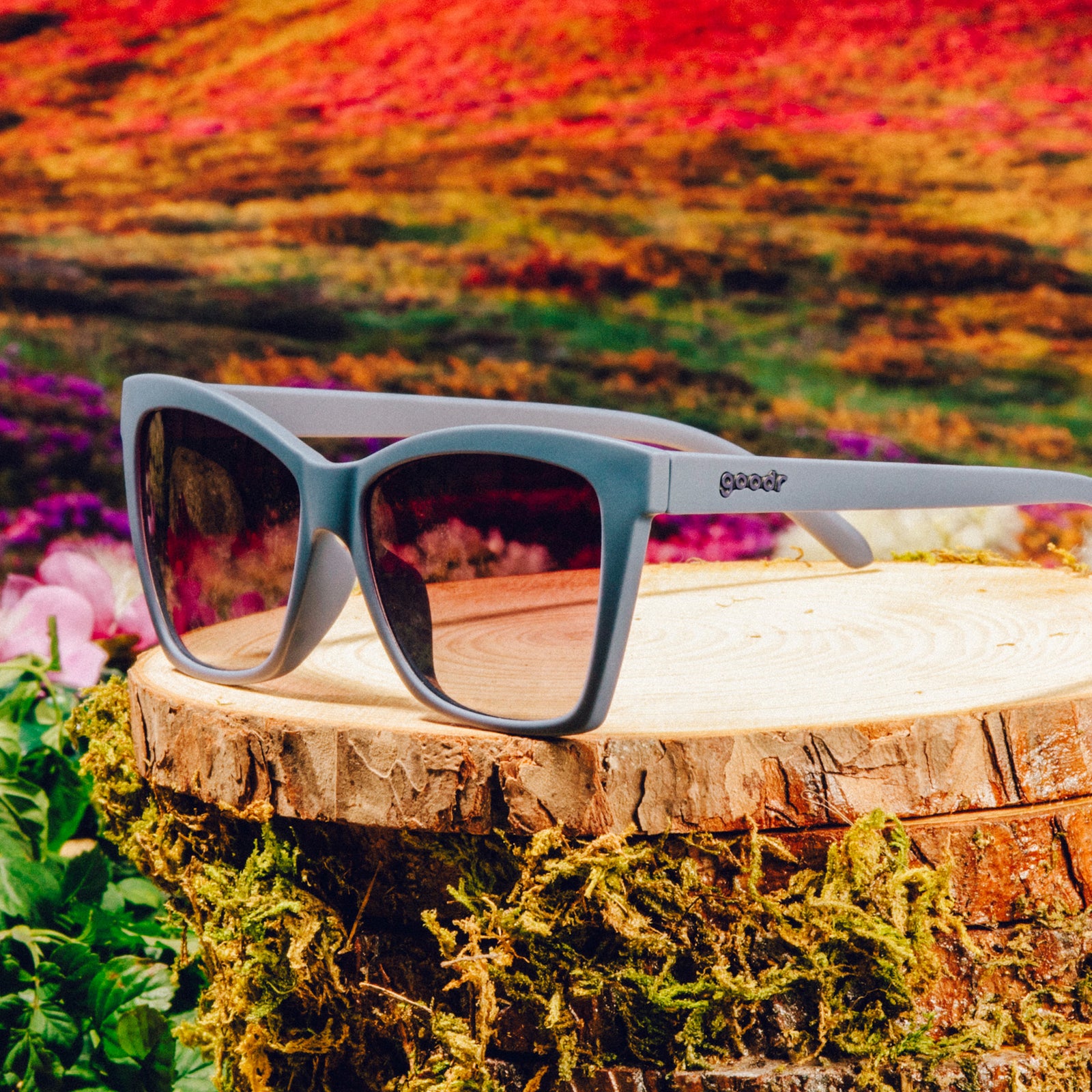 Three-quarter angle view of dark purple angular sunglasses with purple gradient lenses in front of a meadow backdrop.