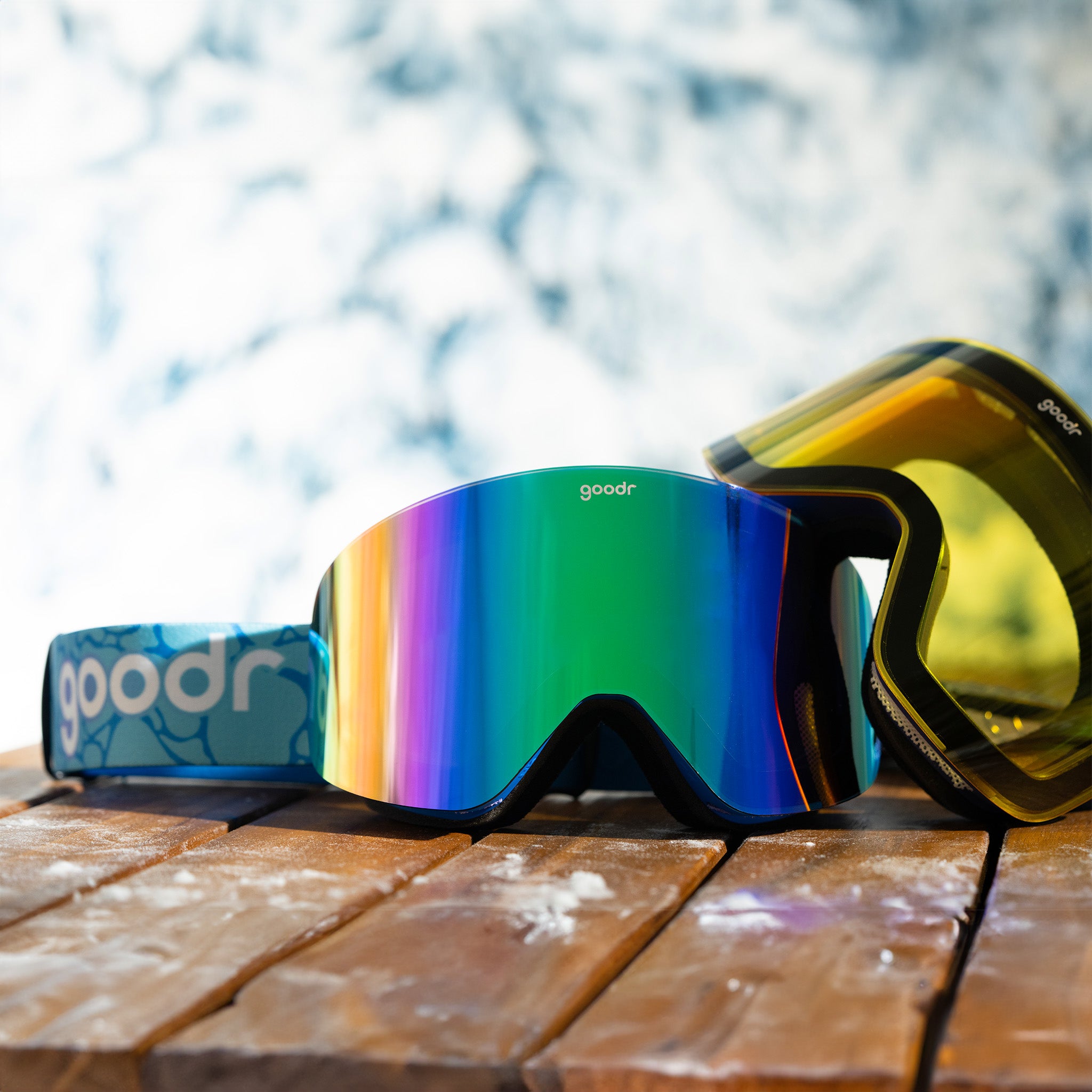 A pair of blue snow goggles rests on a snowy table. A yellow lens meant for low-light conditions is propped up against them.