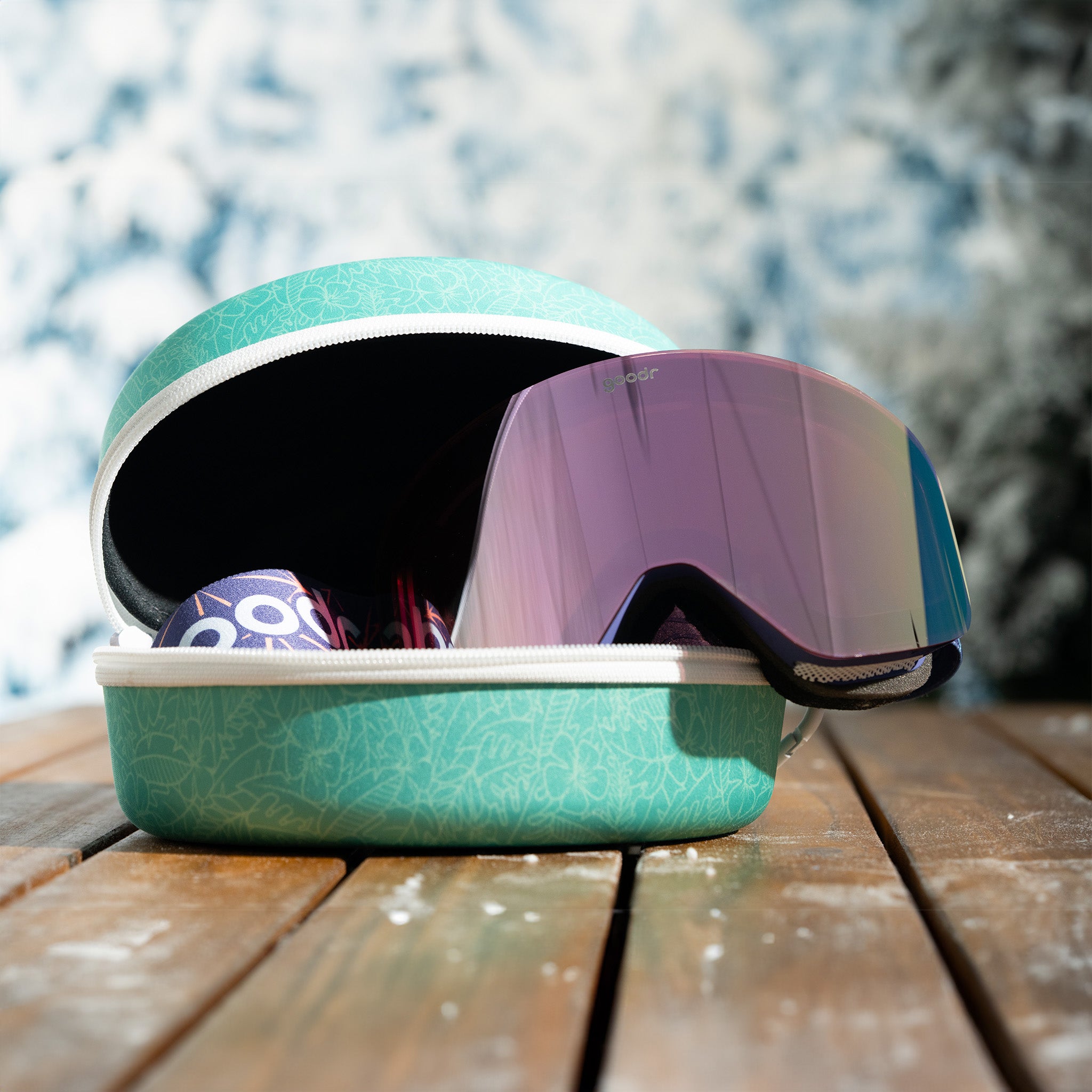 Purple snow goggles are perched inside of a tropical floral print teal zippered case sitting atop a snowy wooden table.