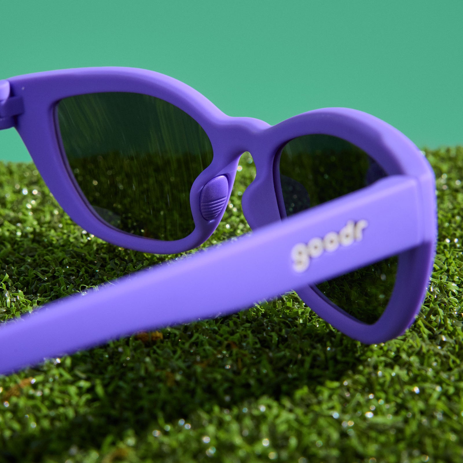 A pair of deep purple cat-eye Cheetah G sunglasses, photographed from behind, sits on astroturf with a mint green background.