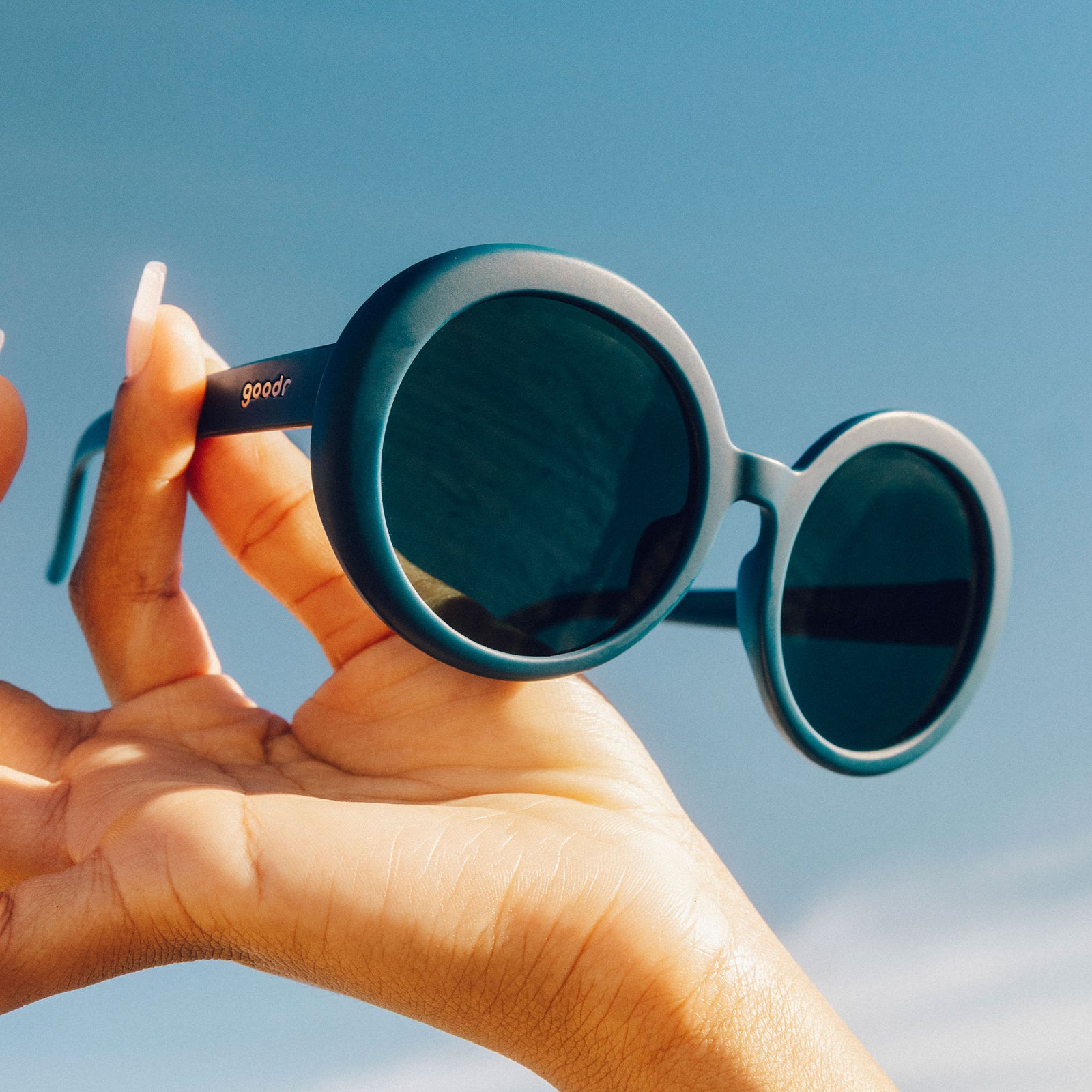 A manicured hand holds up a pair of oversized, round sunglasses in a shade of deep green against the backdrop of a blue sky.