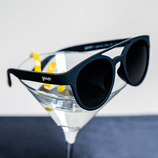 A pair of round, black sunglasses sitting on the rim of a martini glass.