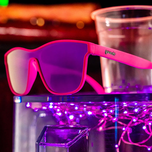 Bright pink, single-frame goodr sunglasses next to a clear plastic cup on a transparent counter top.