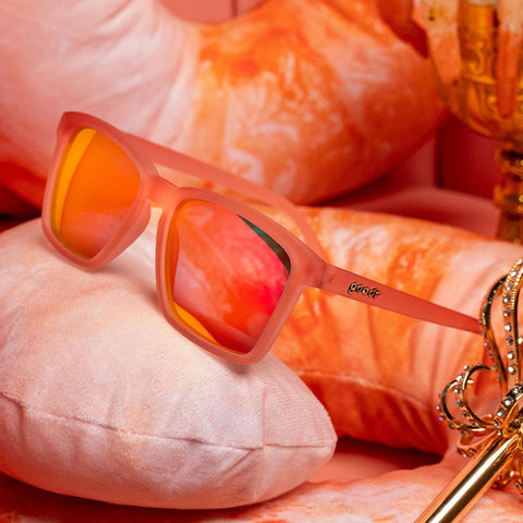 A three-quarter angle view of small square-shaped pink sunglasses with pink mirrored reflective lenses laying on a royal shrimp pillow.