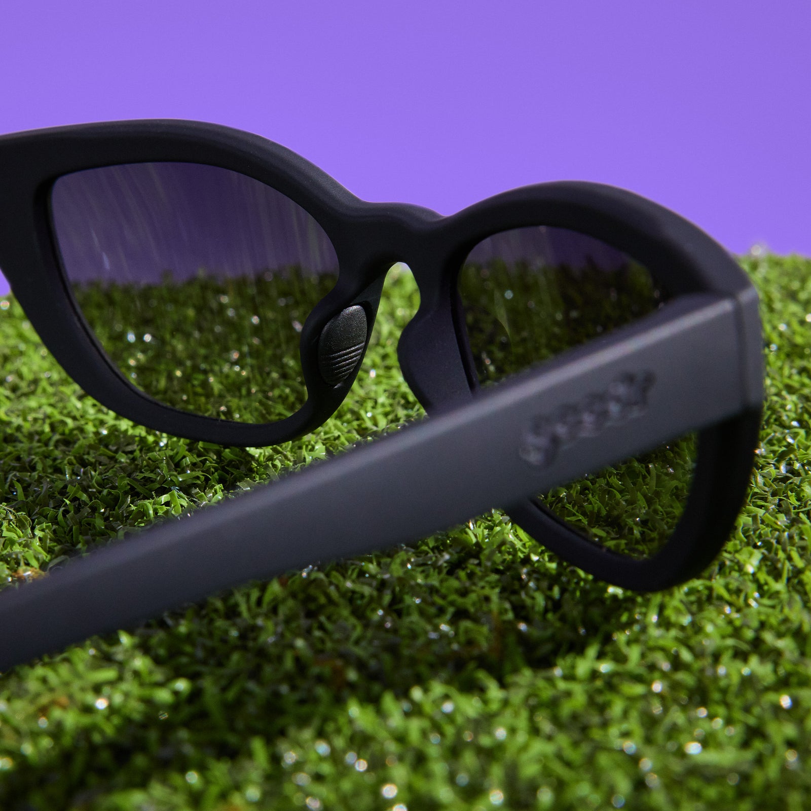 A pair of black cat-eye Cheetah G sunglasses, photographed from behind, sits on astroturf against a purple background.
