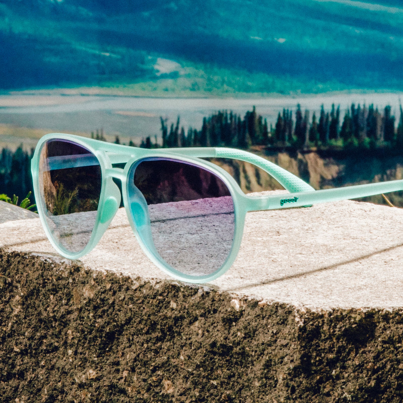Three-quarter angle view of translucent teal aviator sunglasses with dark blue gradient lenses on a boulder against a mountain backdrop.