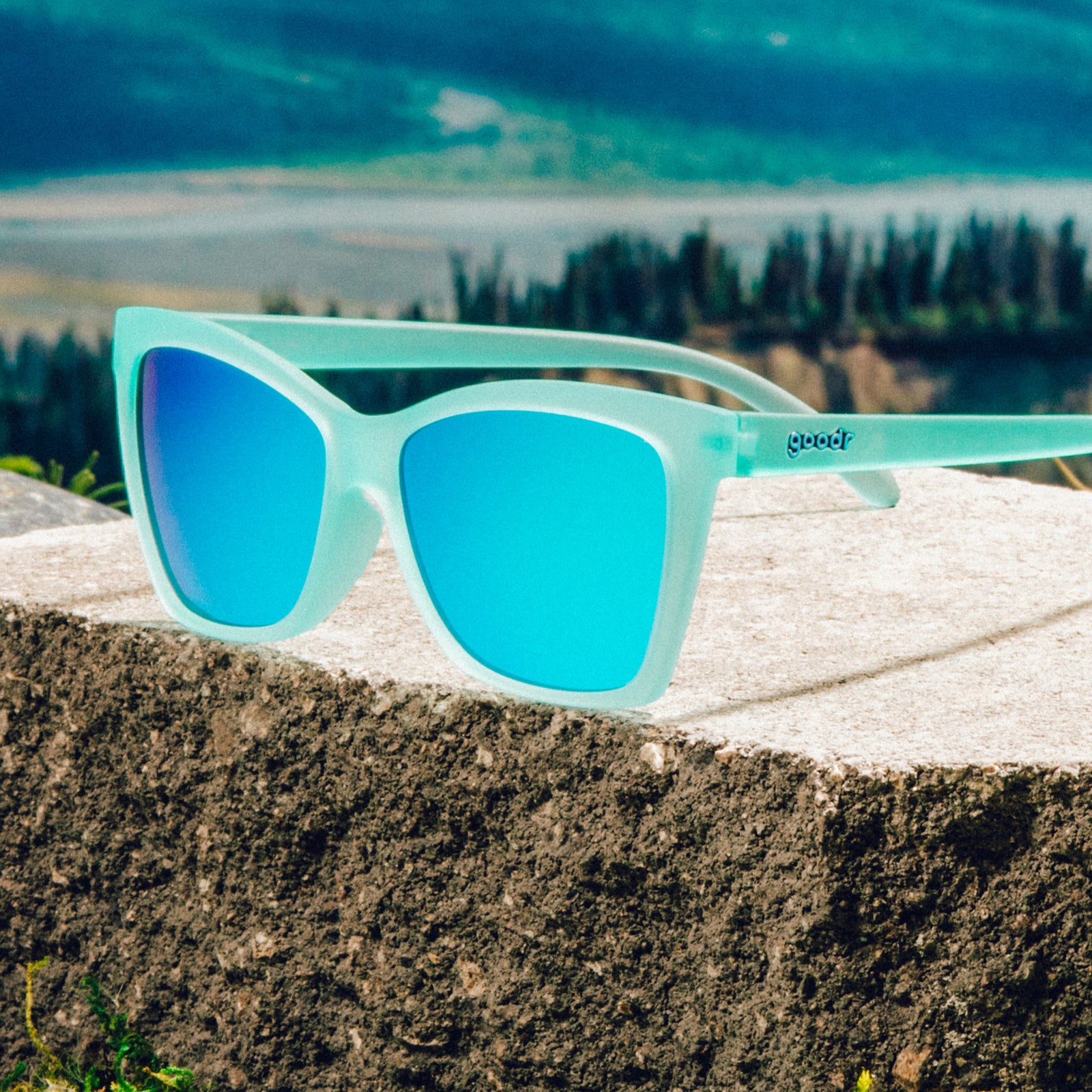 Three-quarter angle view of translucent teal angular sunglasses with reflective teal lenses on a boulder against a mountain backdrop.