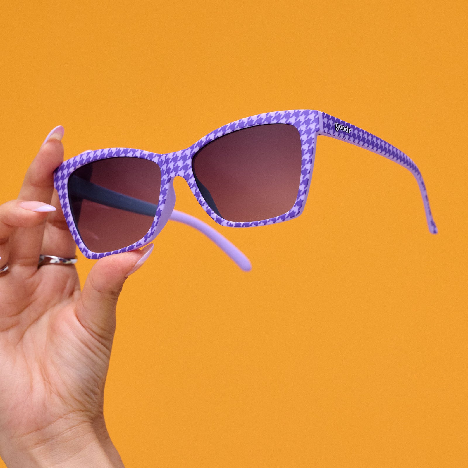 A woman's hand holds a pair of purple houndstooth-print Pop Gs with purple gradient lenses against a mustard backdrop.