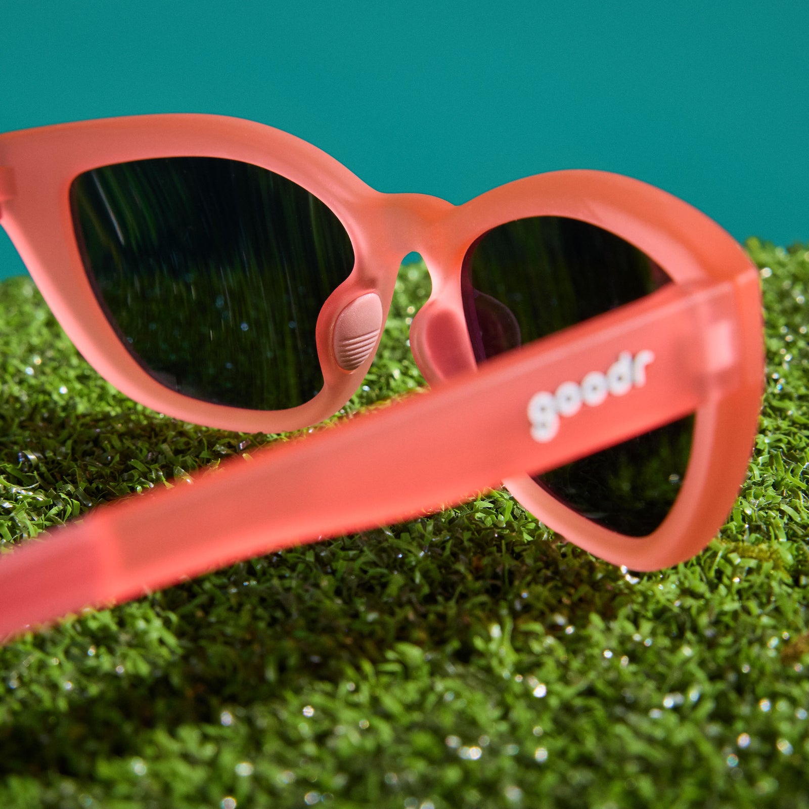 A pair of translucent pink cat-eye Cheetah G frames, photographed from behind, sit on astroturf against a teal backdrop.
