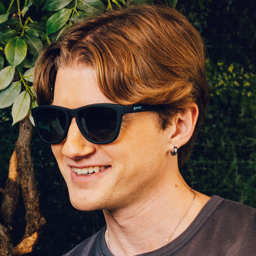 Man smiles while wearing classic dark teal sunglasses with green gradient lenses in front of a nature backdrop.