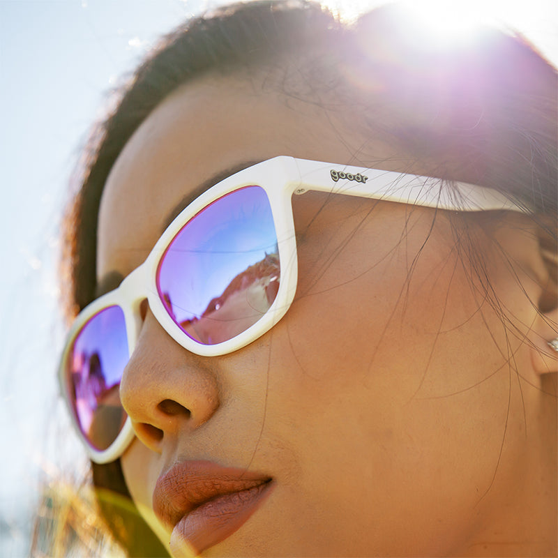 A woman wearing white sunglasses with non-reflective rose-tinted lenses looks off to the side fiercely on a sunny day.